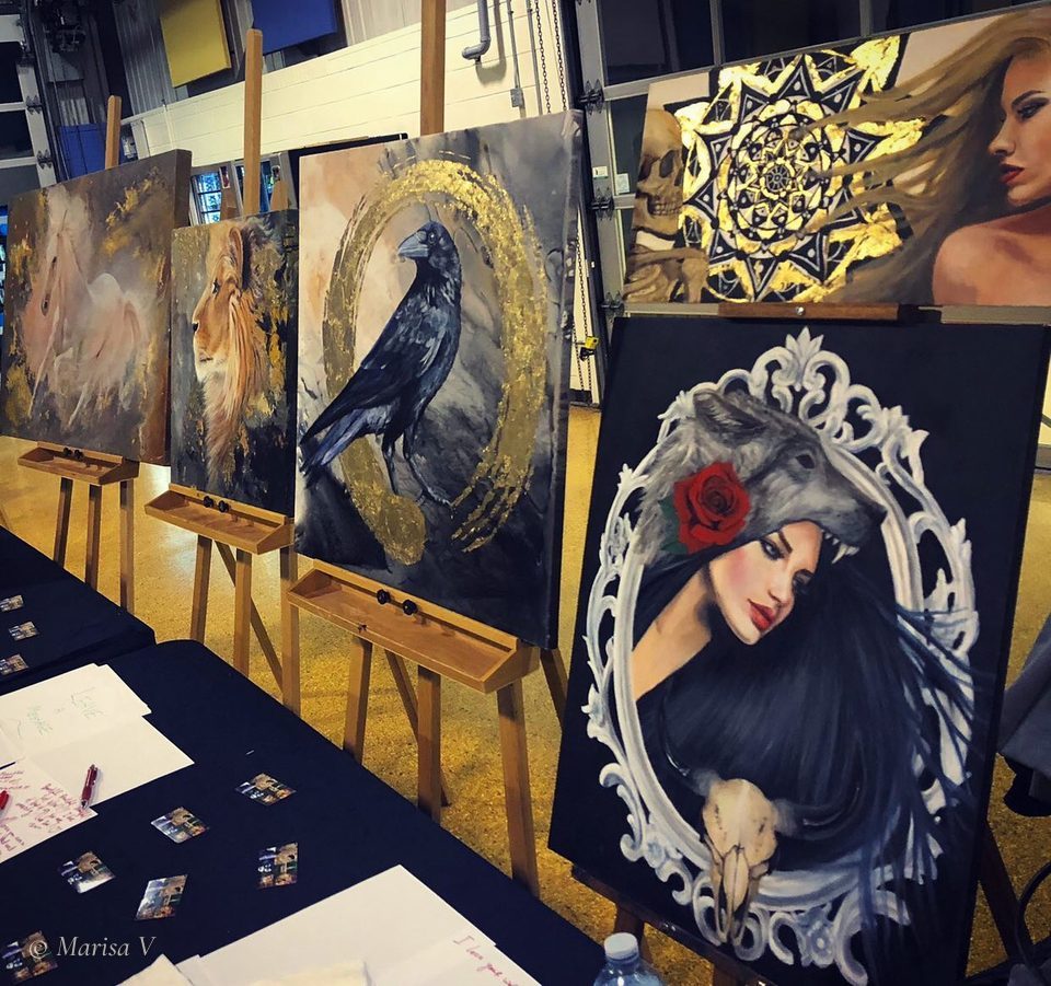 Canvases by Marisa V on easels at an art show: White Pony, Leo, Ensō Circle + Crow, Yellow Eyes and Circle of Life.