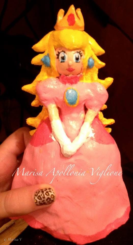 Painted clay figurine of Princess Peach with a gold crown, blonde hair and a pink gown, held in a hand.