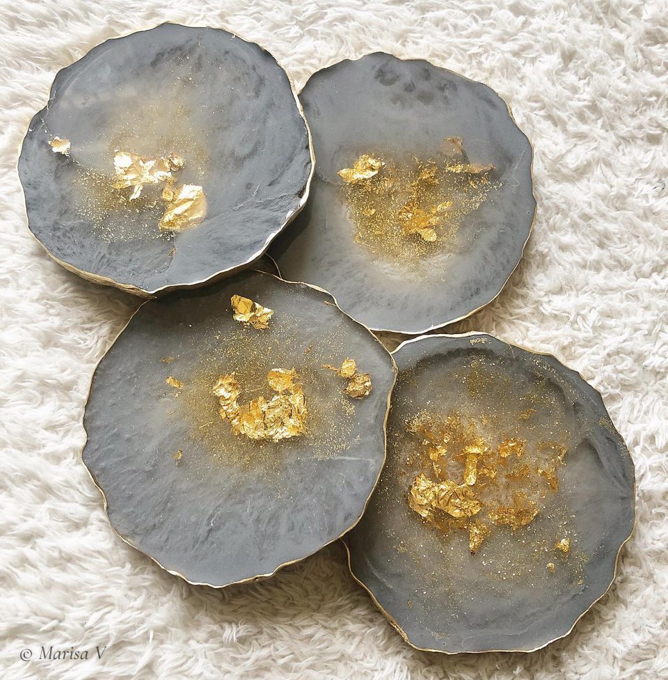 Four smoky grey resin coasters with flakes of gold leaf and gilded rims, on a white rug.