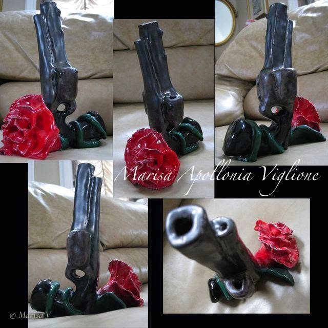 Ceramic sculpture of an upright grey revolver wrapped by a red carnation on a green stem, set on a beige sofa.